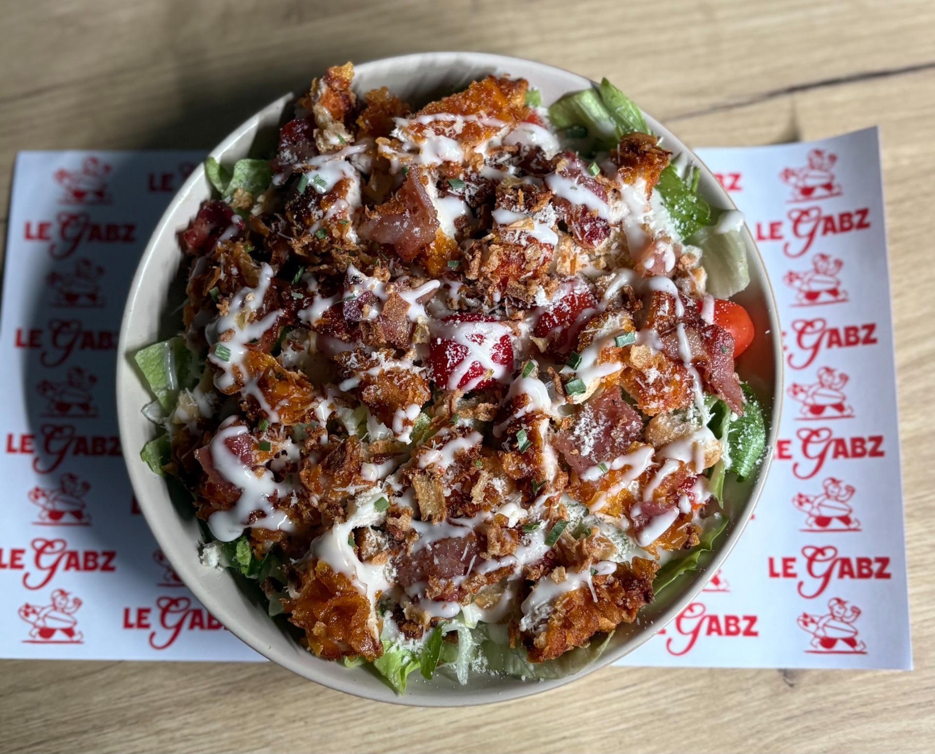La Salade CESAR (Salade fraîche, parmesan, croutons, tomates cerise, poitrine grillée, poulet pané maison, oignons frits, sauce césar)